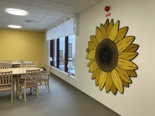 Hörnet av ett rum med matbord och stolar. Den bortre väggen är gul. Väggen till höger är vit och har stora fönster. Bredvid fönstren hänger en stor, gul tuftad matta föreställande en solros. Mattan är skapad av konstnären Jonathan Josefsson.