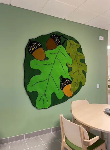 På en grönmålad vägg bakom ett runt bord och stolar, hänger en stor tuftad matta med gröna ekblad i olika nyanser och bruna ekollon. Mattan är skapad av konstnären Jonathan Josefsson.